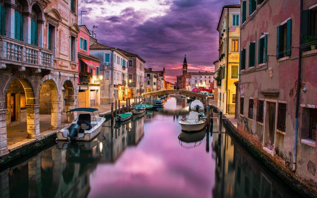 Canal por un paisaje de venecia con tonos mayormente lilas por el cielo y que refleja en el agua las viviendas, donde se muestran también colores vivos de las casas que están situadas a los lados.