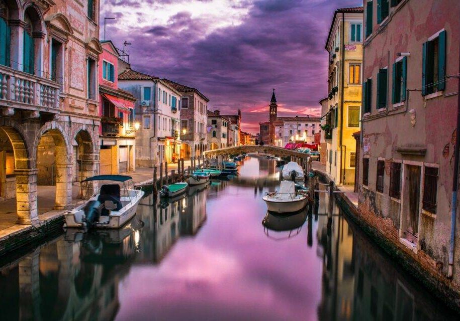 Canal por un paisaje de venecia con tonos mayormente lilas por el cielo y que refleja en el agua las viviendas, donde se muestran también colores vivos de las casas que están situadas a los lados.